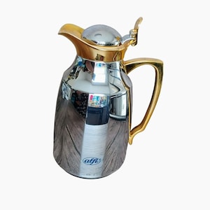 May include: A polished chrome thermal carafe with a gold-colored handle, spout, and lid mechanism. The body features a reflective surface and a blue alfi logo printed near the base. The design includes a hinged lid with a lever for pouring.