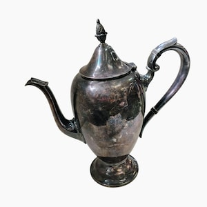 May include: A vintage silver-colored teapot with a curved spout and a decorative handle. The teapot has a lid with a small, ornate finial. The base is a pedestal design. The teapot is likely made of silver or a silver-plated metal.