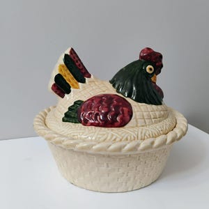 Pode incluir: Um prato de cerâmica em forma de frango com tampa. O frango está sentado numa cesta tecida e é pintado em tons de vermelho, verde, amarelo e branco.