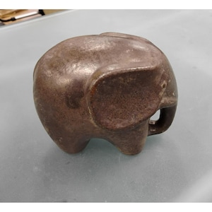 May include: A small, solid, dark brown elephant figurine with a smooth, rounded body, a large ear, and a curved trunk. The surface of the object has a mottled, textured appearance with subtle variations in color throughout the entire form.