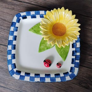 Könnte beinhalten: Ein quadratischer Keramikteller mit weißer Mitte und einem blau-weiß karierten Rand. Eine große, dreidimensionale gelbe Sonnenblume mit grünen Blättern ist auf dem Teller befestigt, begleitet von zwei kleinen roten Marienkäfer-Figuren.