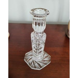 May include: A clear, cut-glass candlestick holder. The candlestick has a hexagonal base and a detailed, textured design. The top is a circular cup to hold a candle. The candlestick is approximately 15 cm tall.