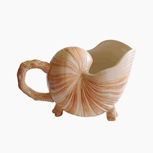 May include: A ceramic pitcher shaped like a nautilus shell, featuring a cream and peach color scheme. The pitcher has a textured handle and rests on three small feet. The shell design includes spiral patterns, creating a unique and decorative piece.