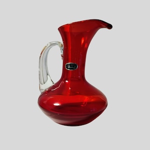 May include: A vibrant red glass pitcher with a clear glass handle. The pitcher has a curved spout and a rounded base. A small, oval label is affixed to the front. The pitcher is likely for serving beverages.