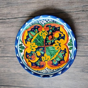 May include: A round ceramic plate featuring a vibrant hand-painted design with a central cross motif. The pattern includes bold yellow scrolls, green leaf-like shapes, and deep blue accents, finished with a blue and white geometric border.