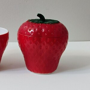 Puede incluir: Un recipiente rojo con forma de fresa con una hoja y un tallo verdes. El recipiente tiene una superficie texturizada con pequeños bultos espaciados uniformemente. La tapa encaja perfectamente y probablemente sea de plástico o cerámica.