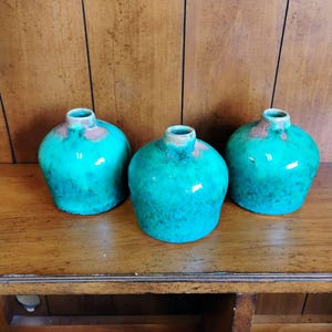 May include: Three turquoise ceramic vases with a glossy finish. The vases have a rounded shape with a narrow neck and a reddish-brown rim. The vases are displayed on a wooden surface.