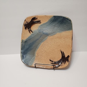 May include: Square ceramic plate with a tan base and a diagonal blue glaze. Two dark brown bird-like figures are on the plate. The plate is displayed on a black metal stand.
