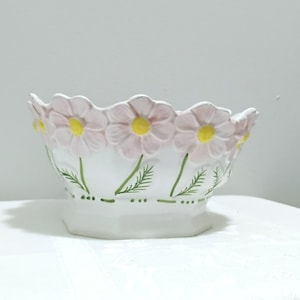 Peut inclure: Un bol en céramique blanche avec un bord festonné en forme de pétales de marguerites rose clair aux centres jaune vif. L'extérieur est orné de tiges et de feuilles peintes en vert, et une ligne verte pointillée entoure la base du bol.