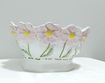 May include: A white ceramic bowl featuring a scalloped rim shaped like light pink daisy petals with bright yellow centers. The exterior is decorated with green painted stems and leaves, and a dotted green line encircles the base of the bowl.