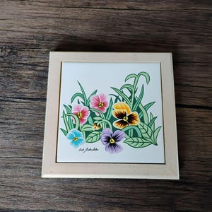 May include: A square ceramic tile featuring a colorful illustration of pansies in shades of pink, yellow, blue, and purple with green foliage, set within a light-colored wooden frame. The artist's signature, Bob Timberlake, is printed in black.