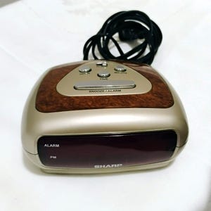 May include: A Sharp brand digital alarm clock with a champagne-colored plastic body and a faux wood grain top panel. It features a dark red display screen, a long black power cord, and silver buttons labeled Hour, Minute, Time, and Snooze/Alarm.