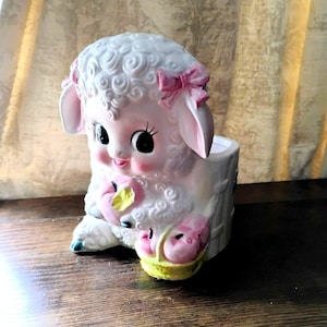 May include: A ceramic lamb planter with a pink bow, ears, and cheeks. The lamb holds a pink fruit with a yellow leaf in a yellow basket. The planter has a white tree trunk design and a hollow interior.