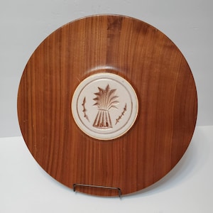 May include: Round wooden serving tray with a central ceramic medallion. The medallion features a wheat sheaf design in brown and cream, set against the warm tones of the wood. The tray is supported by a black metal stand.