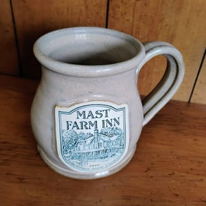 Puede incluir: Una taza de cerámica beige con cuerpo redondeado y asa grande. La taza presenta una etiqueta con el texto "MAST FARM INN" y una ilustración detallada de un edificio y árboles. La taza es probablemente hecha a mano.