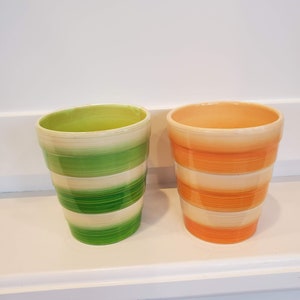 Puede incluir: Dos macetas de cerámica con diseño a rayas. Una maceta es verde y blanca, la otra naranja y blanca. Las macetas tienen forma cilíndrica con un borde ligeramente ensanchado. Están sobre una estantería blanca.