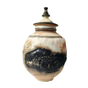 Puede incluir: Un jarrón de cerámica hecho a mano con tapa. El jarrón presenta una superficie texturizada con una paleta de colores crema, negro y marrón. La tapa tiene un pequeño remate decorativo. El diseño del jarrón incluye una banda oscura y texturizada en el medio.