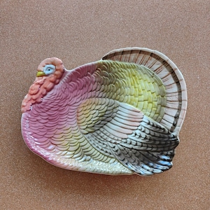 Puede incluir: Una bandeja de cerámica con forma de pavo, que presenta una superficie texturizada con un degradado de plumas rosas, amarillas y marrones. La cabeza del pavo es rosa con un pequeño pico amarillo y un ojo azul, mientras que las plumas de la cola están desplegadas.