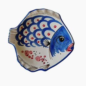 May include: A ceramic fish-shaped plate with a white base, adorned with blue and red painted details. The fish features blue scales, a blue head, and a red mouth. The plate has a blue border and measures approximately 10 inches in length.
