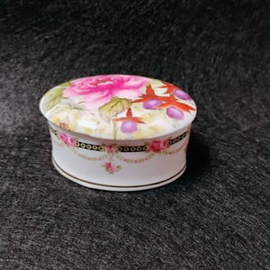 May include: A small, round porcelain trinket box with a floral design. The lid features a large pink rose and other flowers. The box has a white base with a black band and gold trim. A delicate floral garland decorates the base.