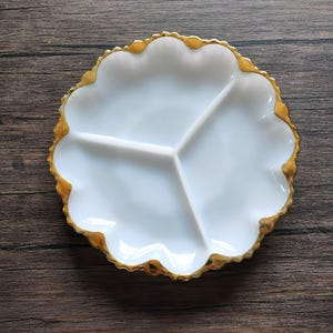 May include: A white, three-sectioned serving dish with a scalloped edge and a gold-colored trim. The dish is designed in a flower shape and is set against a dark wood grain background. Ideal for serving appetizers or snacks.