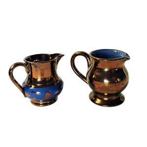 Könnte beinhalten: Zwei kleine, dekorative Krüge mit metallisch bronzefarbenem Finish. Ein Krug hat ein blaues Band mit goldenen Punkten, der andere hat ein blaues Innenleben. Beide haben gebogene Griffe und Ausgüsse, ideal zum Servieren.