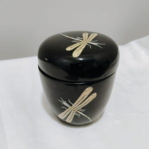 May include: A small, cylindrical black container with a matching lid, featuring a glossy finish. The lid and the body are each decorated with a single, detailed dragonfly illustration in a light, textured beige color with fine crackle details.