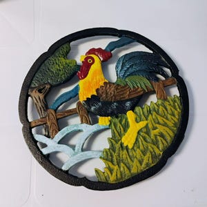 Könnte beinhalten: Wanddekoration aus Gusseisen mit einem Hahn. Der Hahn mit gelbem Körper, rotem Kamm und blauen und grünen Federn sitzt auf einem Holzzaun. Der Hintergrund zeigt einen Baum und Gras, alles in einem schwarzen kreisförmigen Rahmen.