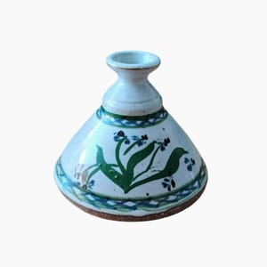 Puede incluir: Un jarrón de cerámica hecho a mano con un esmalte azul claro. El jarrón presenta una base acampanada, un cuerpo cónico y un cuello estrecho. Está decorado con motivos florales y de hojas verdes, y patrones geométricos azules y verdes. La base es de color marrón oscuro.