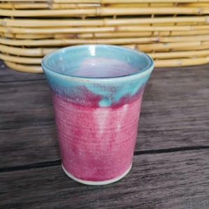 May include: A ceramic tumbler featuring a vibrant pink base that transitions into a smooth turquoise glaze at the rim. The cup has a cylindrical shape with a slightly flared opening and a visible white base, showcasing a handcrafted finish.