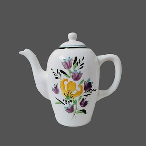 Puede incluir: Tetera de cerámica blanca con tapa, decorada con un diseño floral con flores amarillas y moradas, hojas verdes y detalles en negro. La tetera tiene un pico curvo y un asa redondeada.