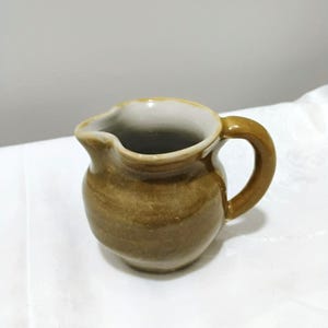May include: A small ceramic creamer jug with a rounded body and a single handle. The exterior features a glossy, mottled brown glaze with horizontal variations in tone, while the interior is finished in a smooth, solid off-white glaze.