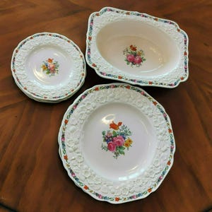 May include: A collection of antique white porcelain dishes with floral center designs. The set includes plates and a serving dish, all with embossed floral borders and colorful floral centerpieces. The dishes are arranged on a dark wooden surface.