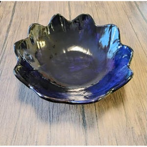 May include: A dark blue ceramic bowl with a scalloped edge. The bowl has a glossy finish and reflects light, showing shades of blue and purple. The bowl is sitting on a wooden surface.