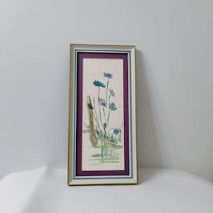 May include: A framed watercolor painting featuring blue flowers and a wooden fence. The artwork is set against a light background with a purple mat and a gold and white frame. The painting evokes a serene, natural theme.