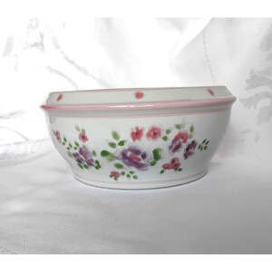 Peut inclure: Un bol en céramique blanche orné d'un motif floral peint à la main avec des touches de rose, de violet et de vert. Le bol présente un bord lisse et arrondi souligné par une fine bande rose et de petits points roses régulièrement espacés.
