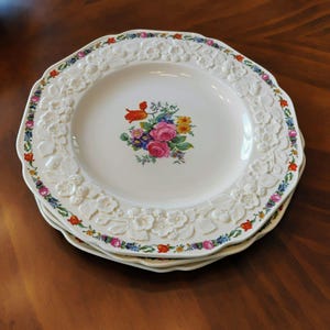 May include: A stack of white ceramic plates with a floral design. Each plate features a raised floral border and a central bouquet of pink, red, and yellow flowers. The plates are stacked on a dark wooden surface.