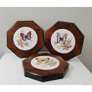 Pode incluir: Conjunto de três bases de madeira com azulejos de cerâmica com desenhos de borboletas. As borboletas são pintadas em tons de azul, vermelho e amarelo com detalhes marrons. As bases são de madeira castanho escura.