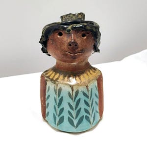 May include: A ceramic figurine shaped like a person with a brown face, dark hair, and a light blue torso decorated with a repeating dark green leaf pattern. The figure has a yellow collar detail and a smooth, glazed finish on the body.