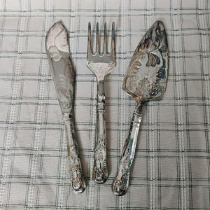 May include: A silver-toned serving set consisting of a knife, a four-tined fork, and a triangular cake server. Each piece features an ornate, embossed floral pattern on the handle and a decorative etched design on the metal surface.