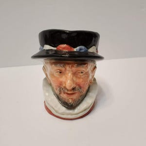Puede incluir: Una jarra de cerámica con forma de cabeza que lleva un sombrero negro con detalles en rojo y azul. La figura presenta un rostro detallado con barba, bigote y un cuello de volantes blanco. La base de la jarra está pintada de color rojo.