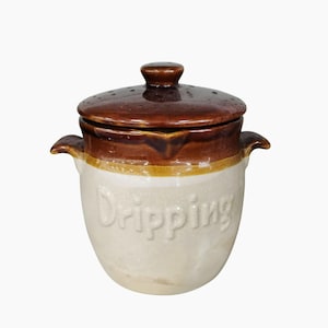 Puede incluir: Una olla de goteo de cerámica vintage con tapa y detalles marrones. La olla tiene una base de color crema con la palabra "Dripping" en relieve en la parte delantera. Cuenta con dos asas laterales y un pequeño pomo en la tapa.