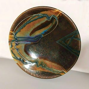 Könnte beinhalten: Eine runde Keramikschale mit einer glänzenden Glasur auf dunkelbraunem Grund. Die Oberfläche zeigt wirbelnde, marmorierte Muster in Blaugrün, Blau und Hellbraun sowie ein markantes, eingraviertes Dreiecksmuster auf der rechten Seite.