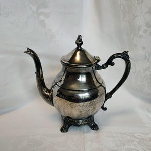 May include: Silver-plated teapot with a curved spout and ornate handle. The teapot has a rounded body, a lid with a decorative finial, and rests on three claw feet. The metal has a polished finish with some visible wear.