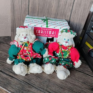 May include: Two plush Christmas dolls with white fur, red noses, and blue eyes, wearing festive outfits. They are dressed in green and red patterned clothing, with matching hats. A box with "Christmas Around the World" text is behind them.