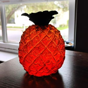 May include: A translucent orange glass pineapple figurine with a textured, diamond-patterned surface and a solid black glass top representing the leaves. The object is illuminated from within, creating a vibrant glow throughout the glass body.