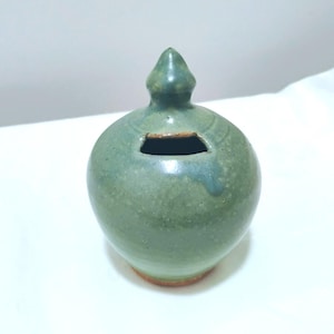May include: A round, sage green ceramic coin bank with a glossy glaze and a pointed lid. The vessel features a horizontal slot at the top for inserting coins, with a visible brown clay base and subtle variations in the green color across the surface.