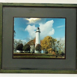 May include: A framed photograph of a white lighthouse with a blue sky and white clouds in the background. The lighthouse is surrounded by trees with green and yellow leaves.