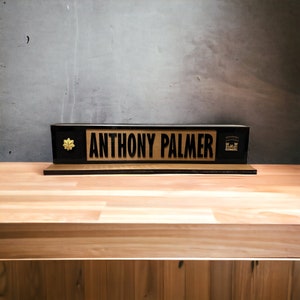 Pode incluir: Uma placa de nome de mesa preta com letras douradas que lê "ANTHONY PALMER". A placa de nome tem uma folha de bordo dourada do lado esquerdo e um emblema dourado do lado direito.