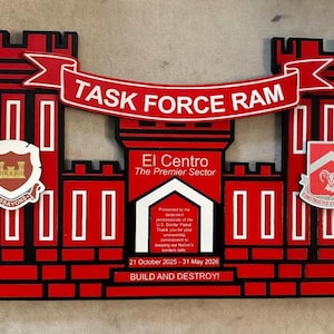 Könnte beinhalten: Rote und schwarze Holzplakette mit der Aufschrift "TASK FORCE RAM" auf einem Banner. Die Plakette zeigt ein Burgdesign mit dem Text "El Centro The Premier Sector" und den Daten "21. Oktober 2025 - 31. Mai 2026".